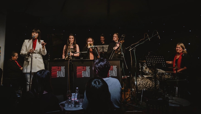 Colour photo of the all female gay big band, with Lara De Belder singing with a band including sax, trumpet, trombone, piano and bass - taken by Jess Rose